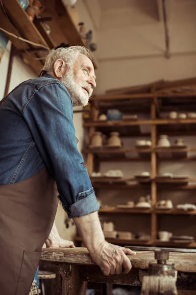 Vista lateral del alfarero senior en delantal de pie y apoyado en la mesa contra los estantes con productos de cerámica en el taller — Foto de Stock