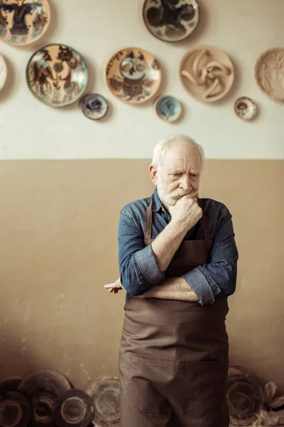 Potier senior dans la pensée tablier et debout contre le mur avec des articles de poterie suspendus — Photo