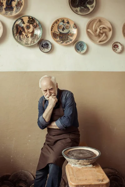 Senior potter in apron thinking and standing against wall with hanging pottery goods — Stock Photo, Image