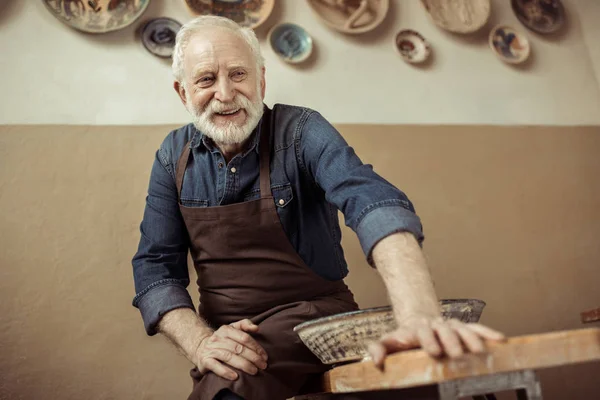 Potier senior dans tablier assis sur la table contre le mur avec des articles de poterie suspendus — Photo