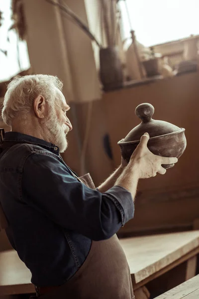 Старший гончар у фартусі, що вивчає керамічну чашу в майстерні — стокове фото