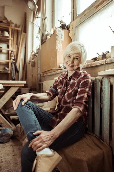 Glückliche Seniorin sitzt in Werkstatt auf Bank neben Fenster — Stockfoto