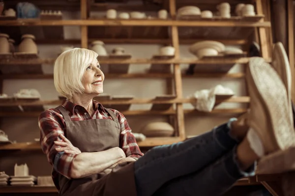 Front view of senior woman sitting on chair with legs on table against shelves with pottery goods — Stock Photo, Image