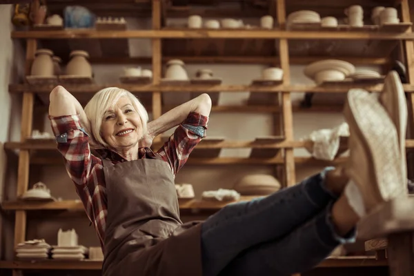 Вид спереди пожилой женщины, сидящей на стуле с ногами на столе на фоне полок с гончарными изделиями — стоковое фото