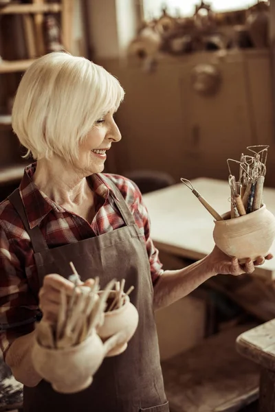 Старша жінка тримає чаші з керамічними інструментами в майстерні — стокове фото