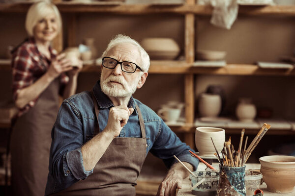 Senior potter sitting at table and thinking with senior woman at workshop
