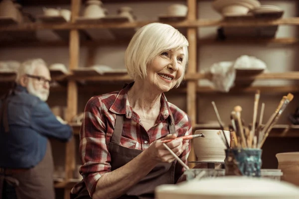 Woman painting clay pot with senior potter at workshop — Stock Photo, Image