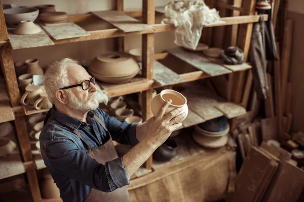 Alfarero senior en delantal y anteojos examinando cuenco de cerámica en el taller —  Fotos de Stock