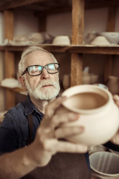 Alfarero senior en delantal y anteojos examinando cuenco de cerámica en el taller — Foto de Stock