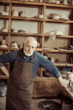Üst düzey potter ayakta ve seramik ürünler ile kamera duvara bakarak önlük