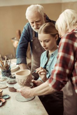 Side view of girl painting clay pot and grandparents helping at workshop clipart