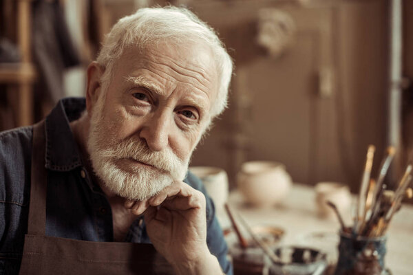 Portrait of senior potter in apron sitting at table and looking at camera