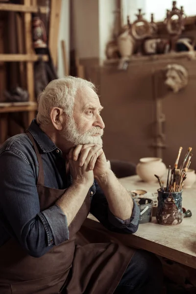 Senior potter in apron sitting at table and daydreaming at manufacturing — Stock Photo, Image