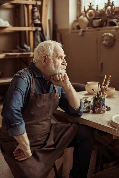 Senior potter in apron sitting at table and daydreaming at manufacturing