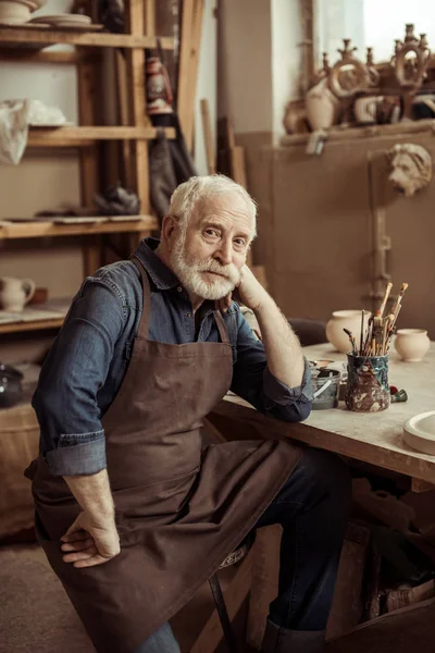 Senior potter i förklädet sitter vid bord och tittar på kameran vid tillverkning — Stockfoto