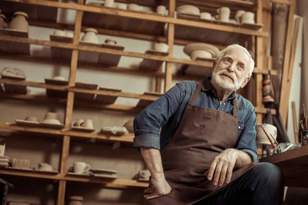 Senior potter i förklädet sitter vid bord och dagdrömma vid tillverkning — Stockfoto