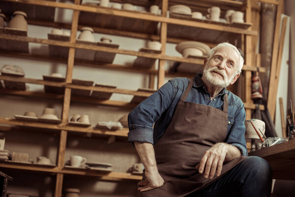 Senior potter in apron sitting at table and daydreaming at manufacturing