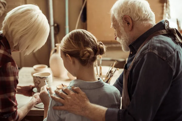 Задний вид девушки, рисующей глиняный горшок, и бабушки и дедушки, помогающие в мастерской — стоковое фото