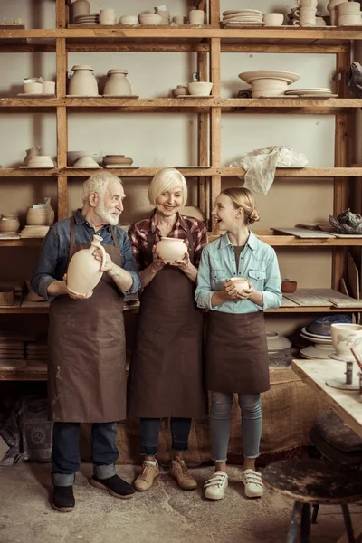 Granddaughter and grandparents standing and holding clay vase and bowls against wall with pottery goods — Stock Photo, Image