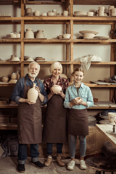 Granddaughter and grandparents standing and holding clay vase and bowls against wall with pottery goods