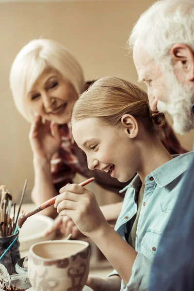 Side view of girl painting clay pot and grandparents helping at workshop — Stock Photo, Image