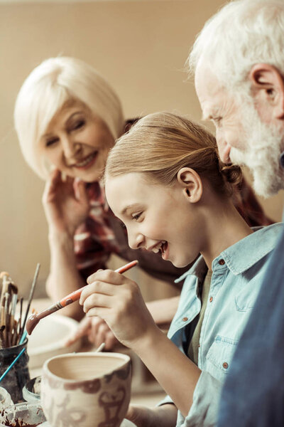 Вид сбоку на девочку, рисующую горшок, и бабушку, помогающую в мастерской
