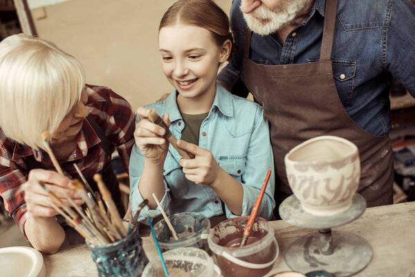 Front view of girl painting clay pot and grandparents helping at workshop