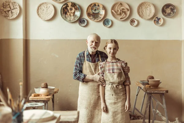 Front view of senior potter with his granddaughter in aprons standing at workshop — Stock Photo, Image