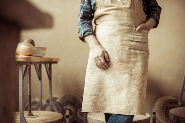 Cropped image of senior potter in apron standing at workshop