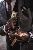 Částečné prohlédnout americký podnikatel držení sklenice alkoholu nápoj a doutník při skrytí bankovky dolaru v kapse saka obleku