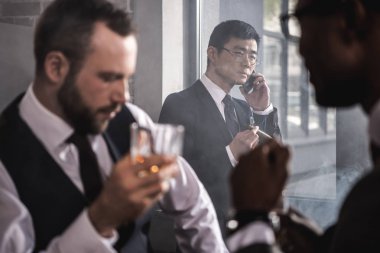 Puro sigara ve viski, çok kültürlü iş takım içme meslektaşları süre smartphone konuşurken ciddi Asya iş adamı