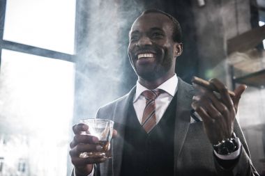 Afrika kökenli Amerikalı işadamı bardak viski ve sigara puro ile tutarak gülümseyen
