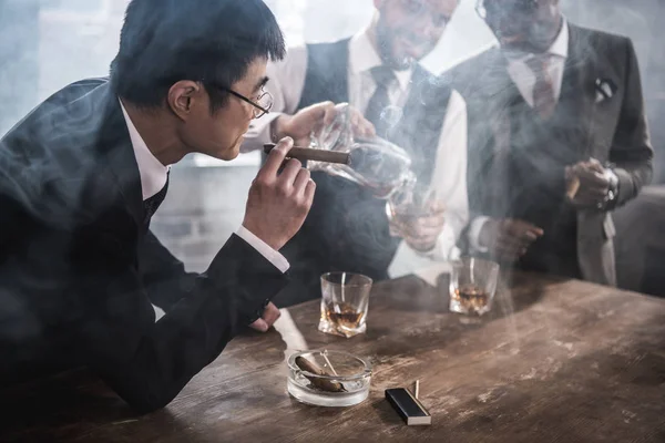 Itsevarma aasialainen liikemies tupakointi sikari ja katsomalla kollegansa juo viskiä — kuvapankkivalokuva