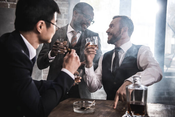 Multiethnic group of businessmen smoking and drinking whisky indoors, multicultural business team