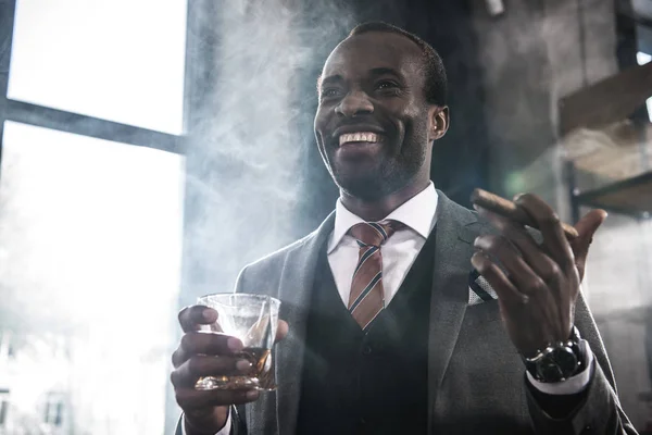 Smiling african american businessman holding glass with whiskey and smoking cigar — Stock Photo, Image