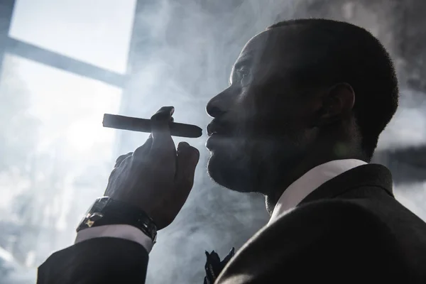 Vista lateral do homem americano africano confiante fumar charuto dentro de casa — Fotografia de Stock