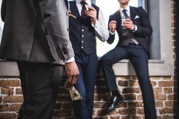 Foto recortada de un hombre de negocios sosteniendo un billete de dólar y mirando a sus colegas bebiendo whisky, reunión del equipo de negocios — Foto de Stock