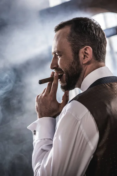 Vista lateral de homem confiante sorrindo fumar charuto dentro de casa — Fotografia de Stock