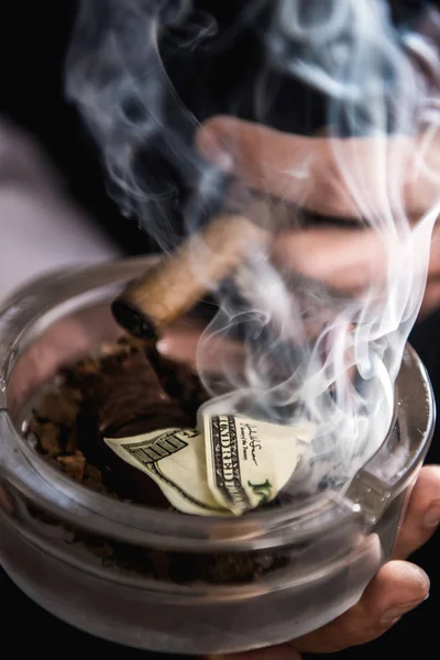 Vista parcial de cerca del hombre fumando cigarro y sosteniendo el cenicero con billete de dólar — Foto de Stock