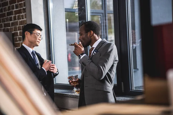 Confident businessman drinking alcohol beverage and smoking cigar while colleague hiding money into suit pocket, business team meeting — Stock Photo, Image