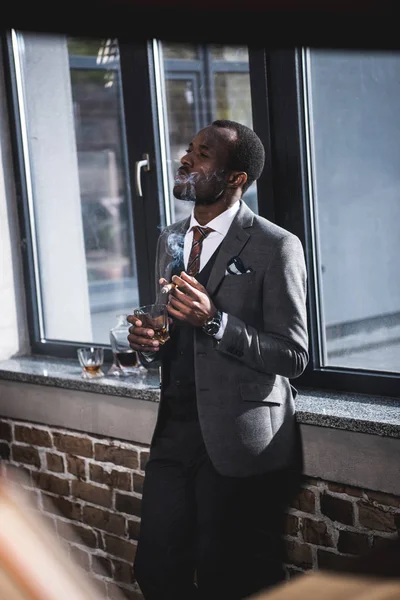 Уверенный бизнесмен держит стакан с виски и курит сигару в помещении — стоковое фото