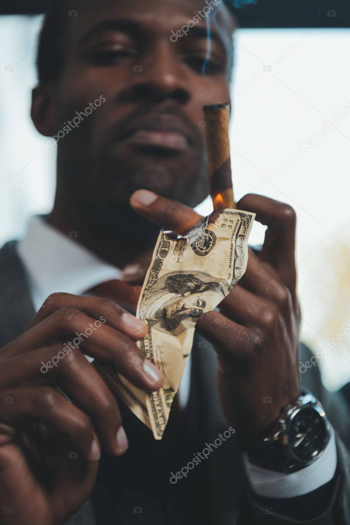 confident african american businessman smoking cigar and burning dollar banknote 