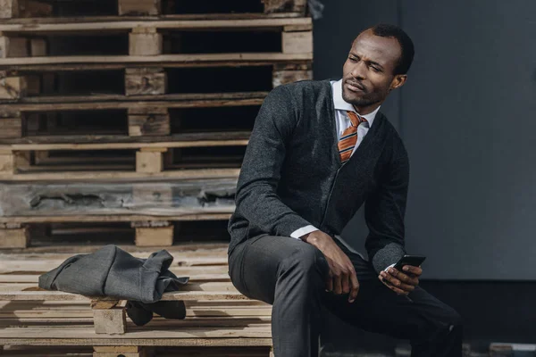 Κουρασμένος κομψό Αφρικανικός Αμερικανός επιχειρηματίας χρησιμοποιώντας smartphone και να κάθονται σε εξωτερικούς χώρους — Φωτογραφία Αρχείου