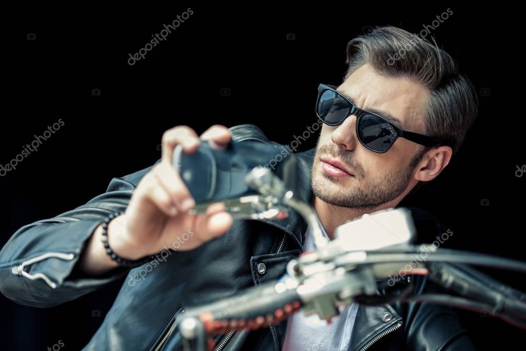 Handsome stylish young man in sunglasses and leather jacket sitting on motorcycle and looking away