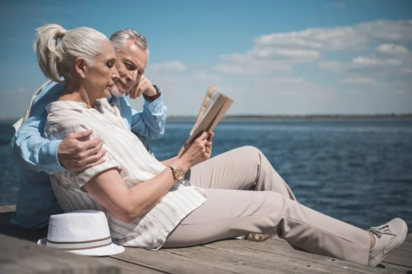 Elderly couple reading book on quay — Stock Photo, Image