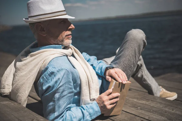 Senior man lying with book on quay — Stock Photo, Image