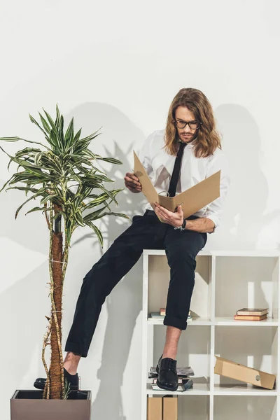 Businessman with folder sitting on bookshelf — Free Stock Photo