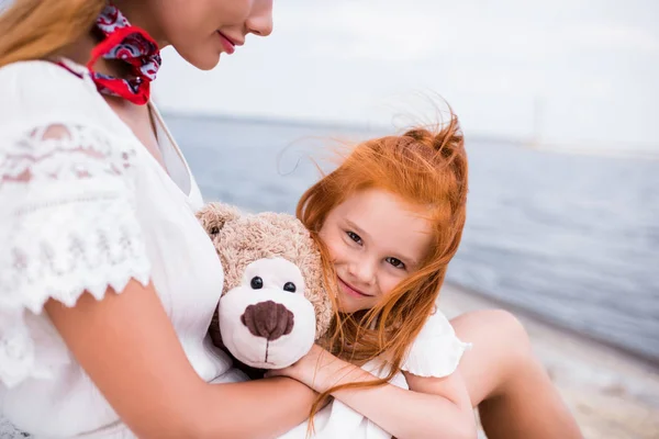 Mother and daughter with teddy bear at seashore — Stock Photo, Image
