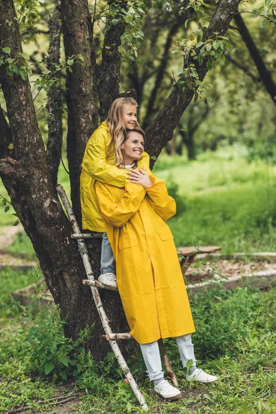 Mother and daughter in raincoats at tree — Stock Photo, Image