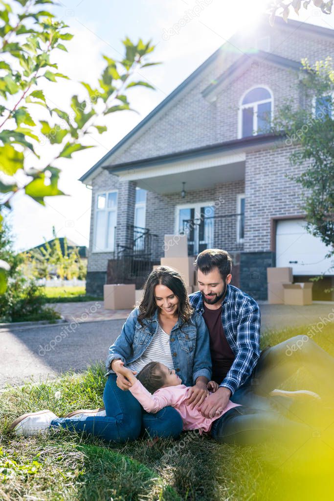 Young parents cuddling adorable little daughter on grass after moving into new house